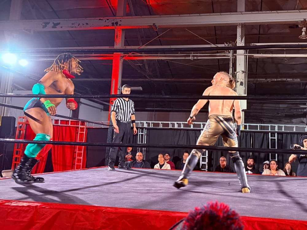 Two wrestlers, seen through the ring ropes, face off with a ref between them.